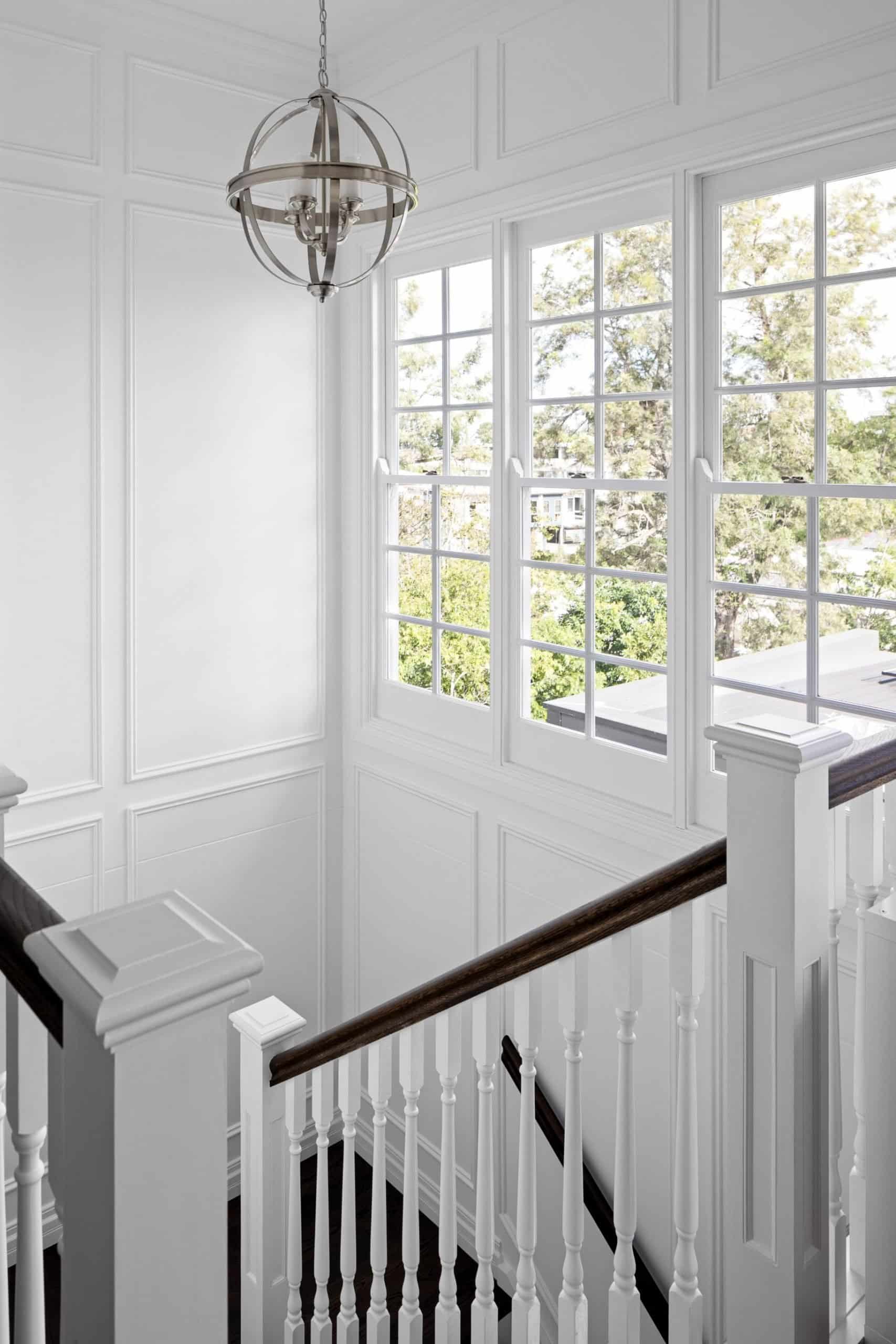 Freshly painted staircase in Brisbane home with white panelled walls, timber handrail and large windows